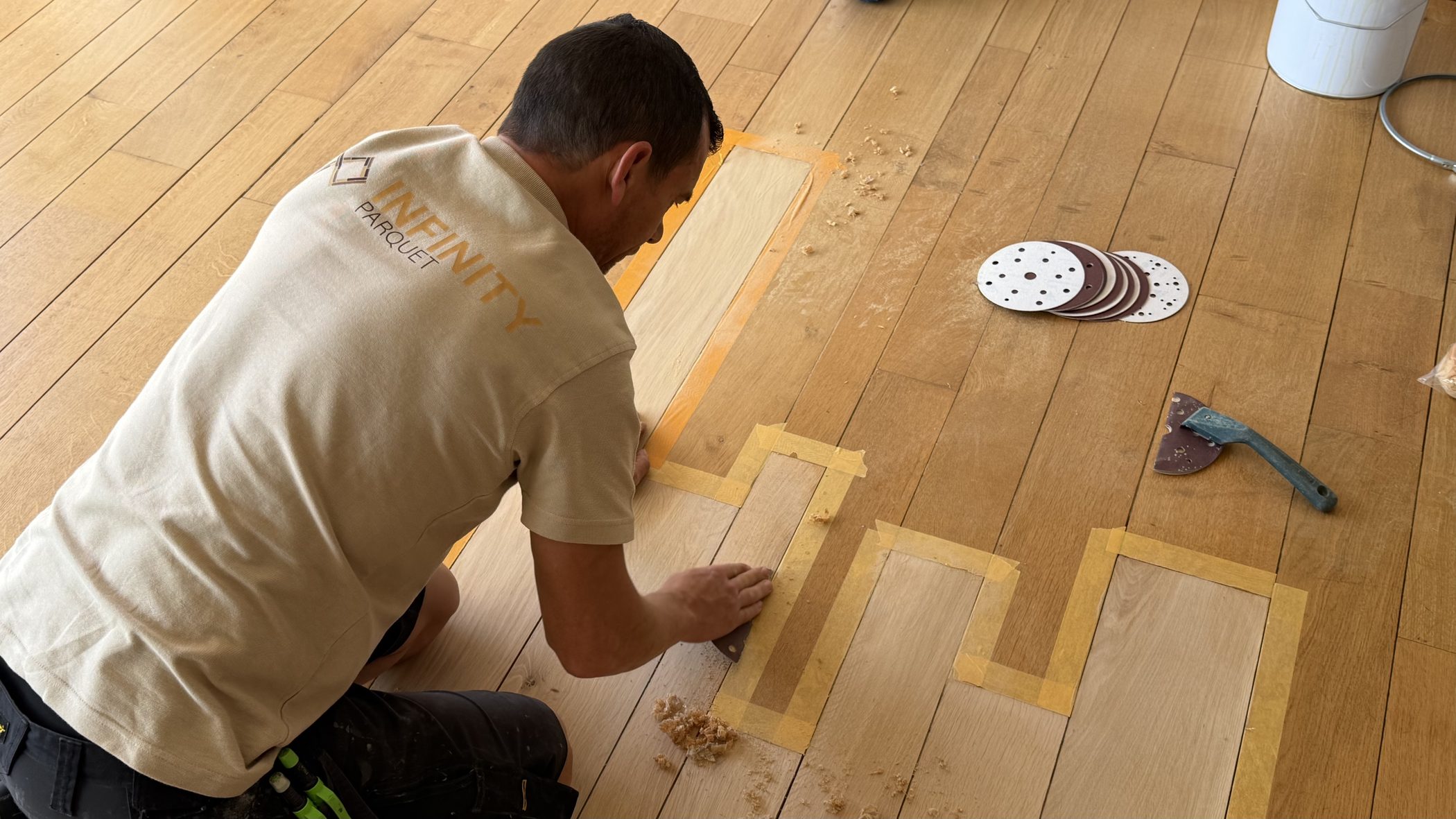 Artisan Infinity Parquet à genoux, en train d'ajuster une pièce de parquet marquée au ruban